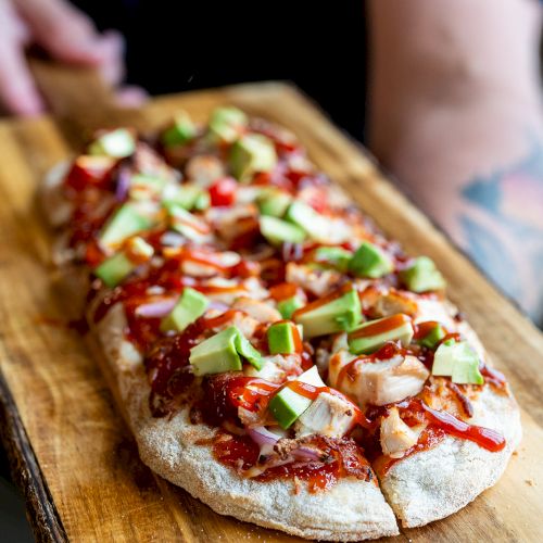 A person holds a wooden board with a rectangular pizza topped with red sauce, chicken, avocado, and onions.